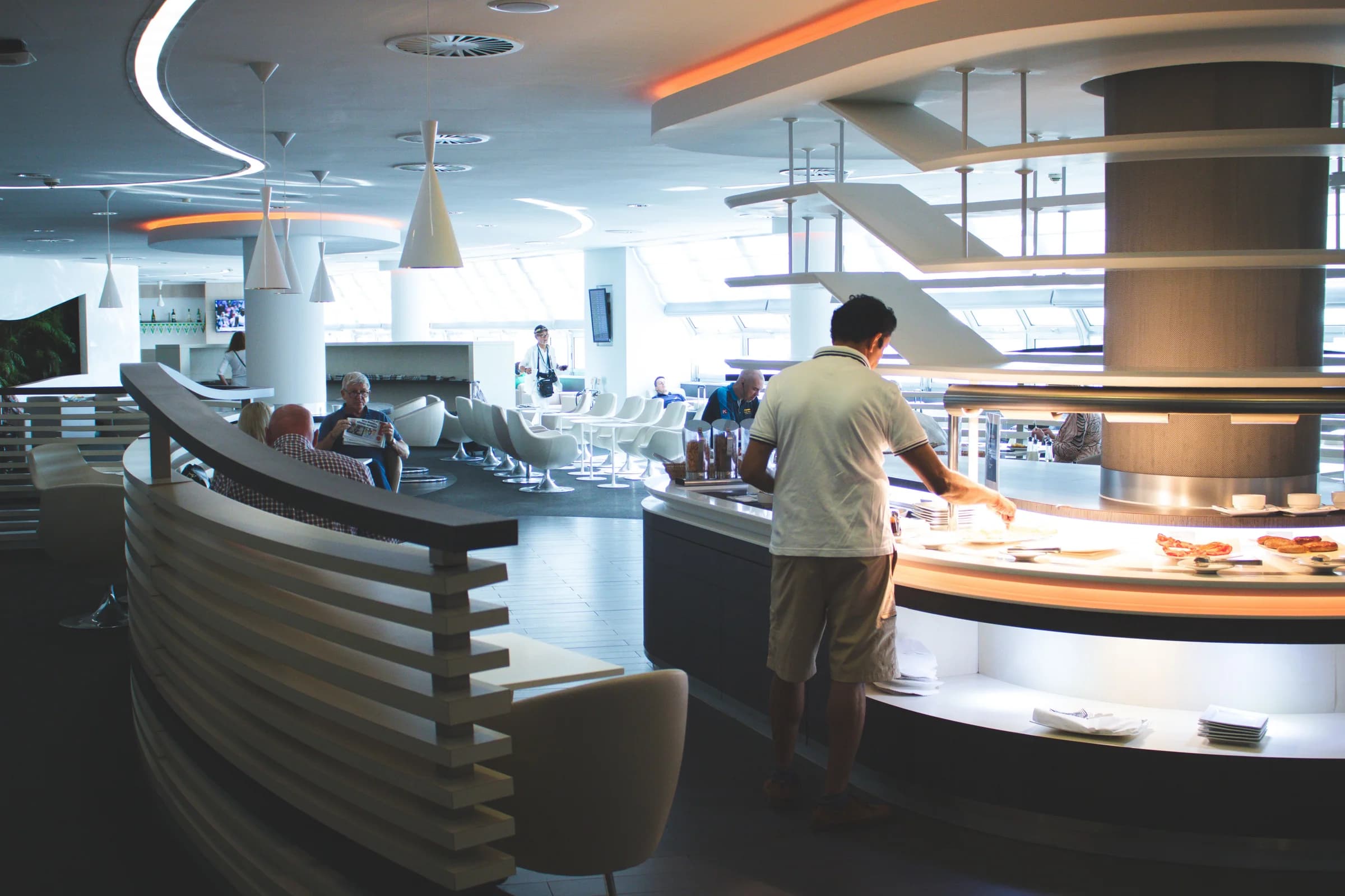 Gäste in einer hellen Flughafen-Lounge, ein Mann bedient sich am Buffet, andere lesen in Sesseln