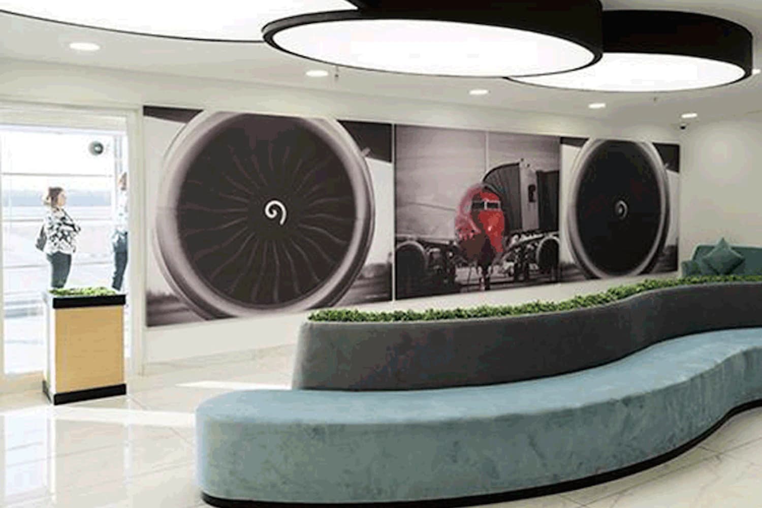 Geschwungene Sitzbank mit Mittelbegrünung vor großem Wandbild zweier Flugzeugtriebwerke und runden Deckenleuchten.