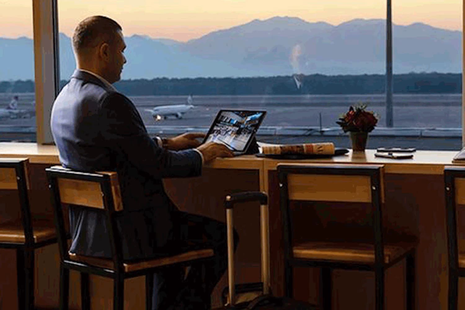 Mann im Anzug sitzt an Fensterbar in Flughafenlounge, arbeitet am Laptop, Trolley neben ihm, Startbahn und Berge im Sonnenuntergang.