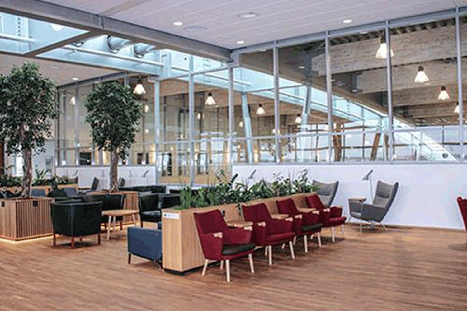 Lounge mit Reihen roter Sessel, einzelnen Sesseln, Holzboden, Pflanzen und Glaswand zum Terminal.