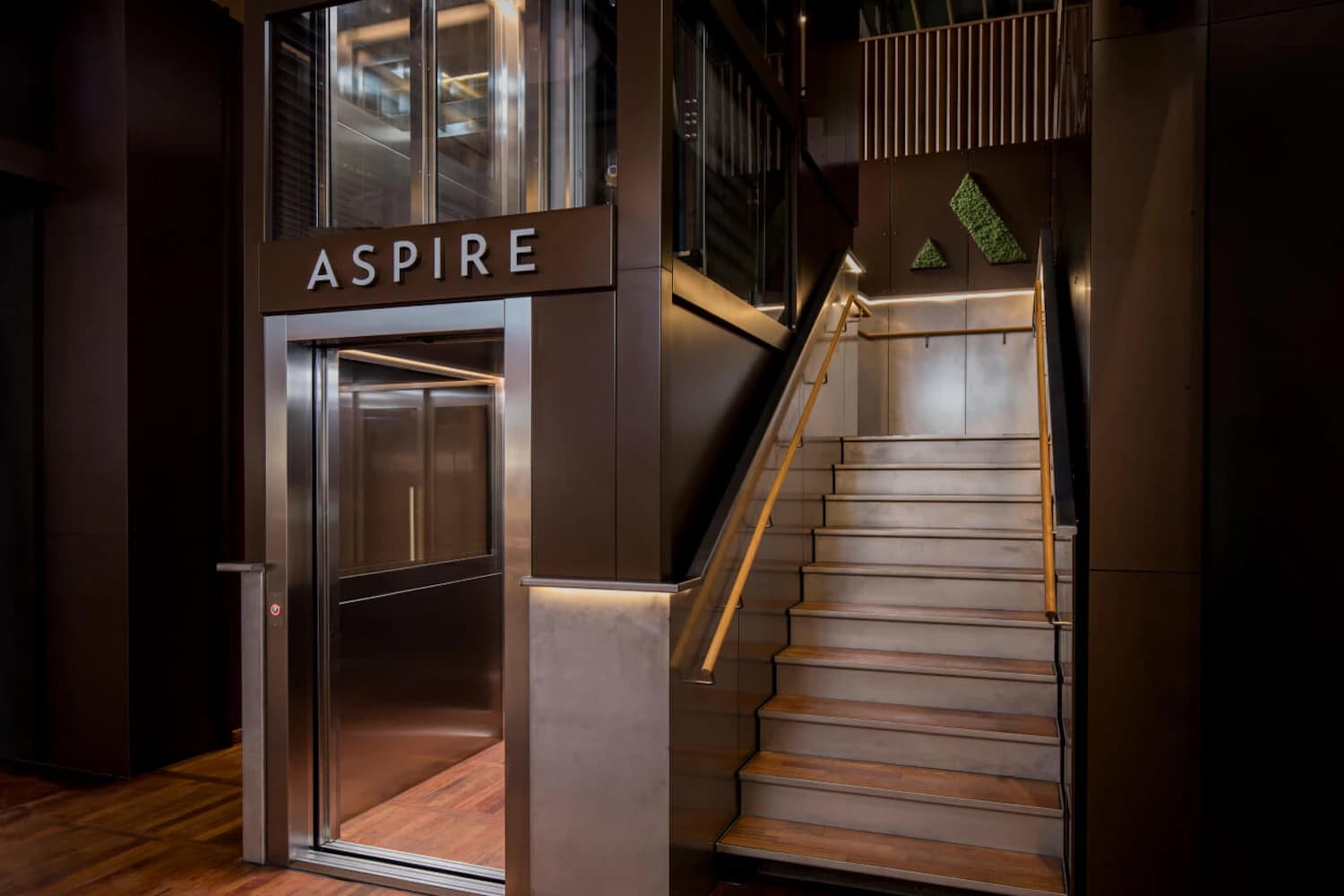 Eingang zur Aspire mit offenem Aufzug, rechts eine Treppe mit Holzstufen und goldfarbenem Handlauf.