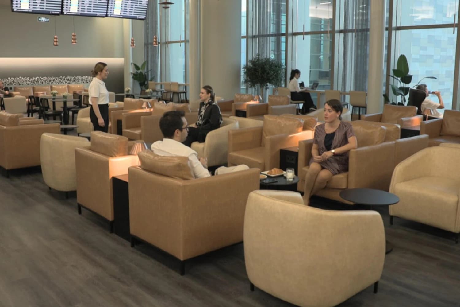 Lounge mit beigen Ledersesseln, Gästen an Tischen, großen Fenstern und Anzeigetafeln im Hintergrund.