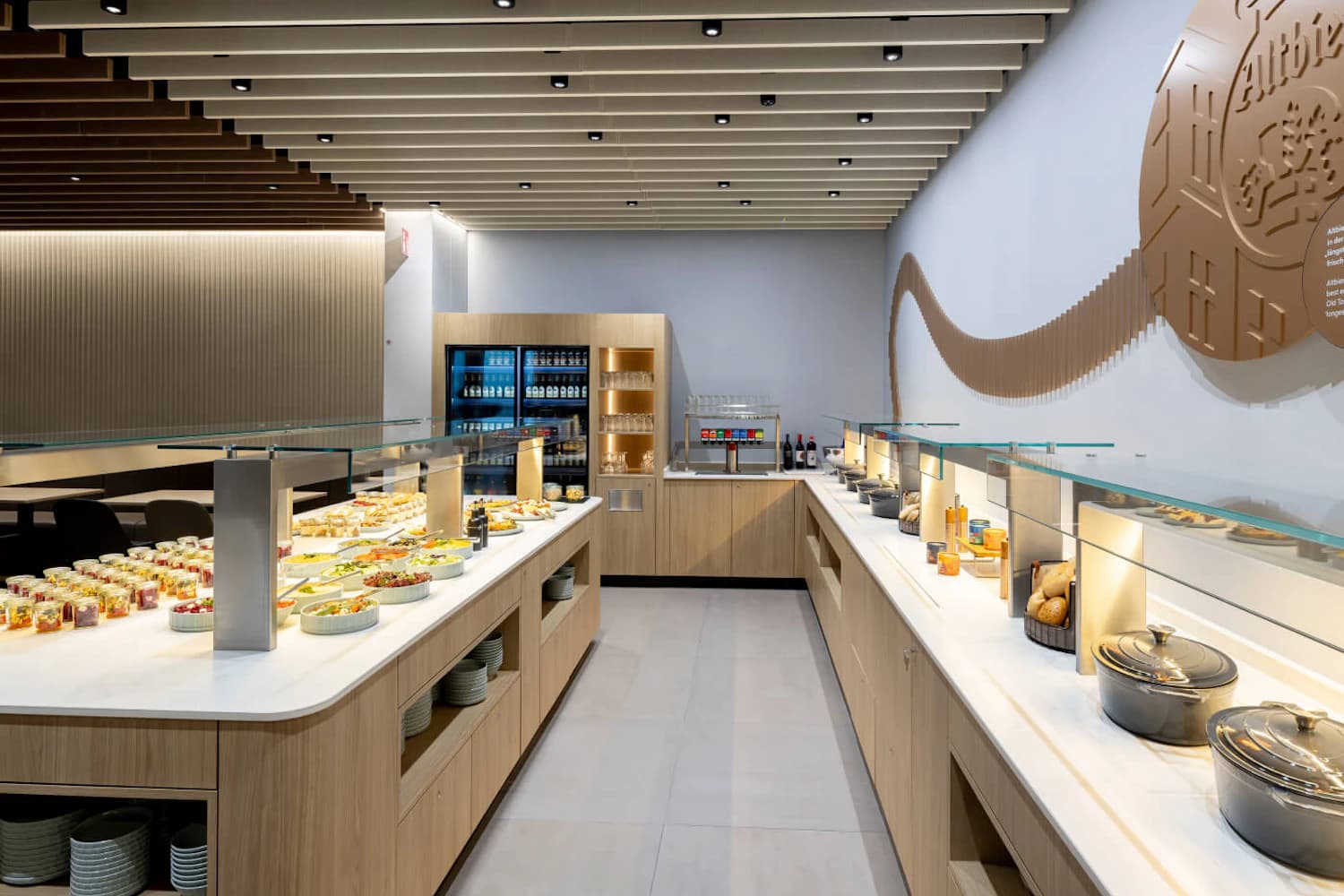 Buffetbereich in heller Lounge mit zwei Servierinseln, Tellern, Getränkekühlschrank und Wanddekoration.