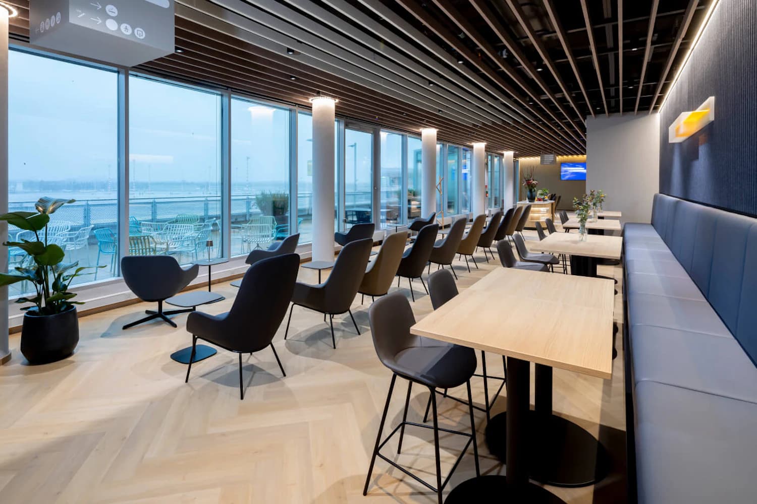 Helle Lounge mit Reihen moderner Sessel und Tische, großen Panoramafenstern zur Außenterrasse und Blick aufs Vorfeld.