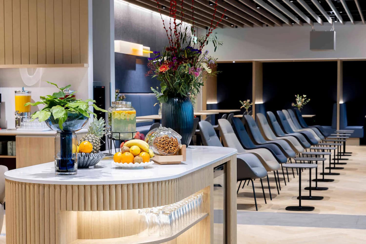 Lounge mit Theke vorn mit Obst, Gebäck und Getränkespender; dahinter Reihen moderner Sessel und kleine Tische.