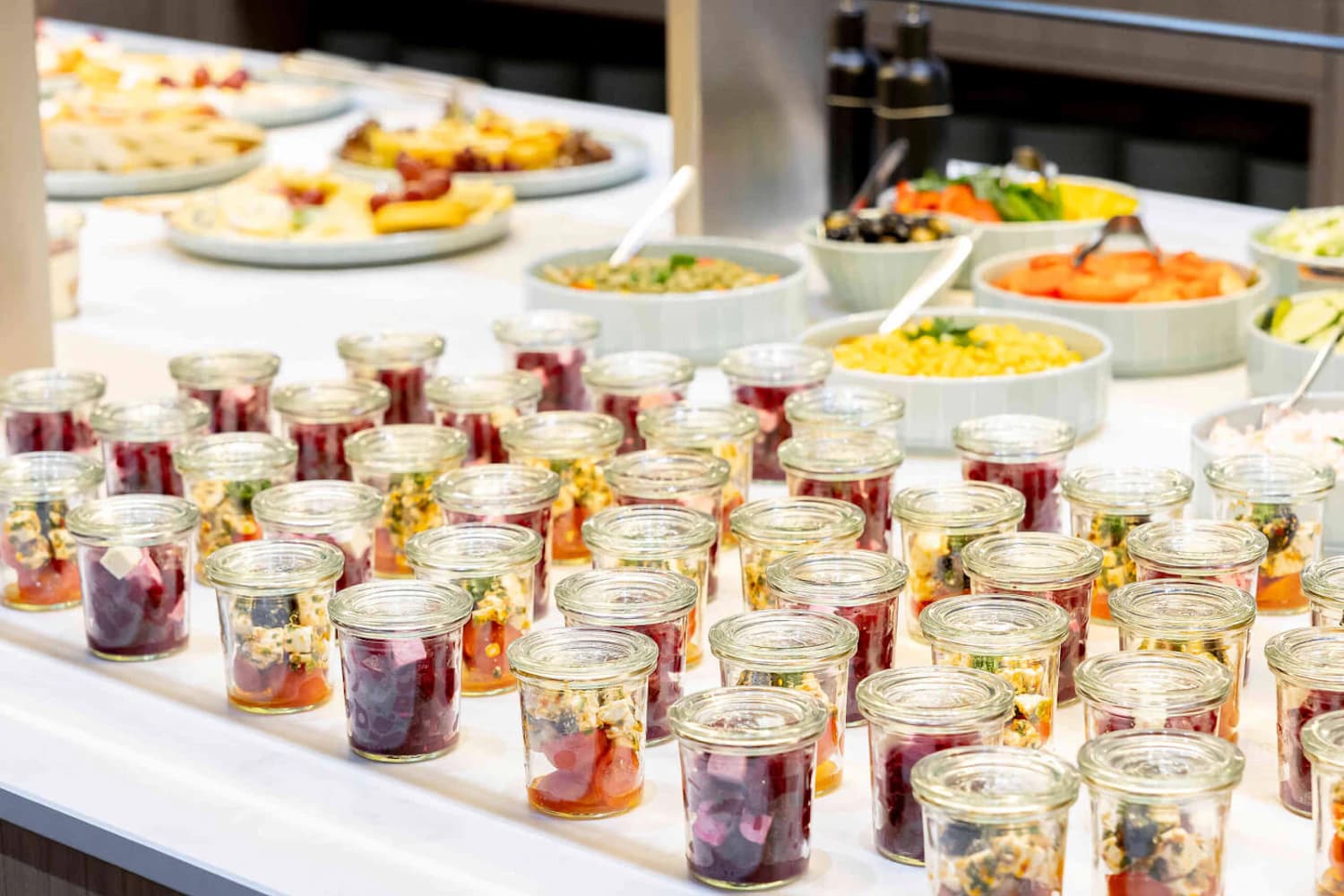 Viele kleine Glasgläser mit Salatportionen auf einem Buffet, im Hintergrund Schalen mit Mais, Gurken und Oliven.