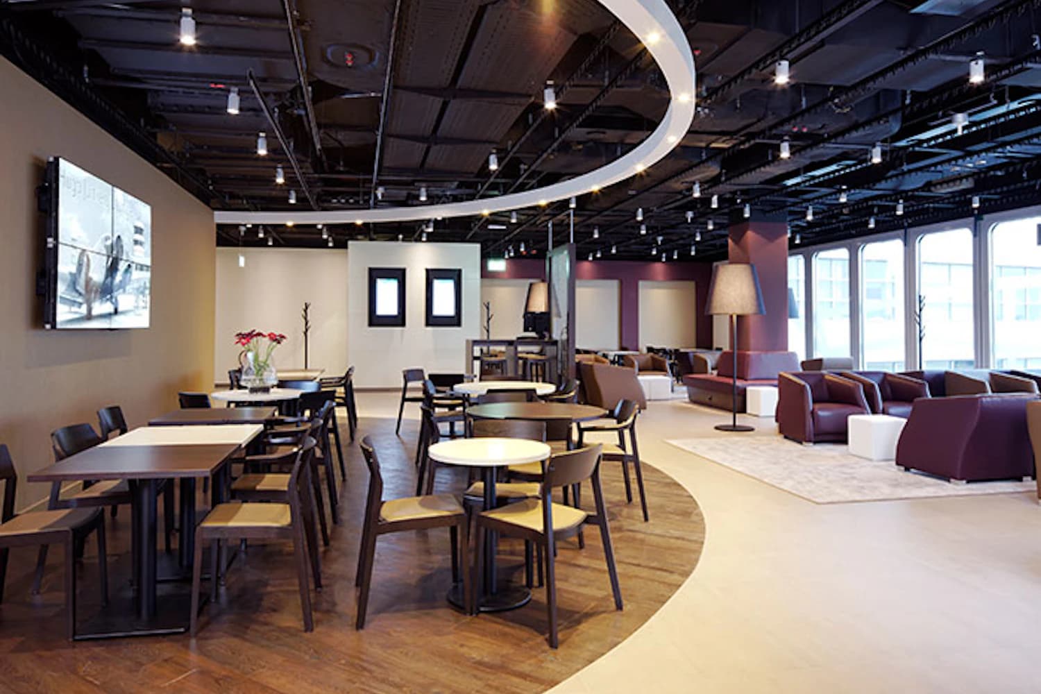 Lounge im Flughafen mit Tischen, Stühlen, Sitzgruppen und geschwungener Deckenbeleuchtung.