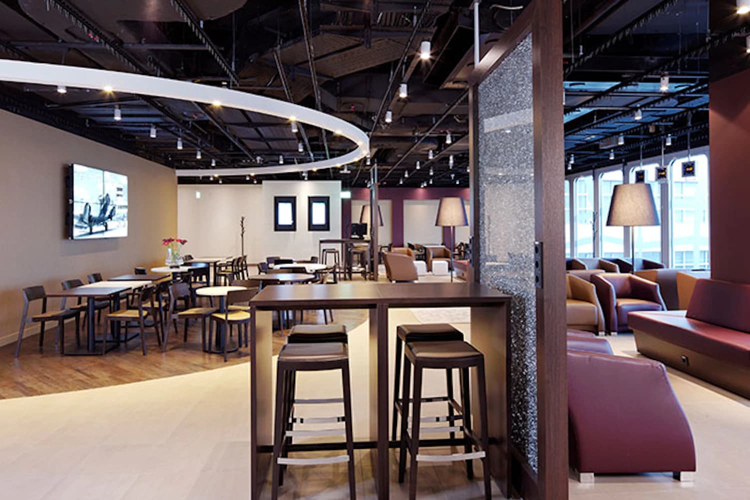 Helle Lounge mit Hochtisch und Hockern, Tischen, Sesseln, Stehlampen und rundem Deckenlicht.