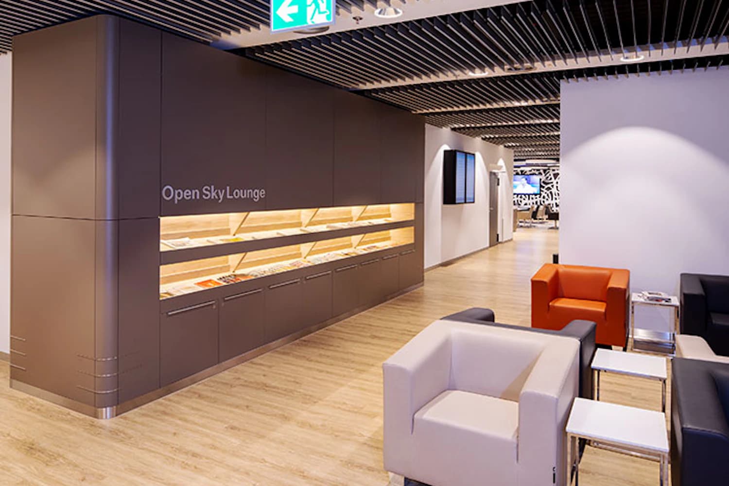 Helle Flughafenlounge mit Sitzgruppen, beleuchtetem Magazinregal beschriftet Open Sky Lounge und Fluchtwegschild an der Decke.
