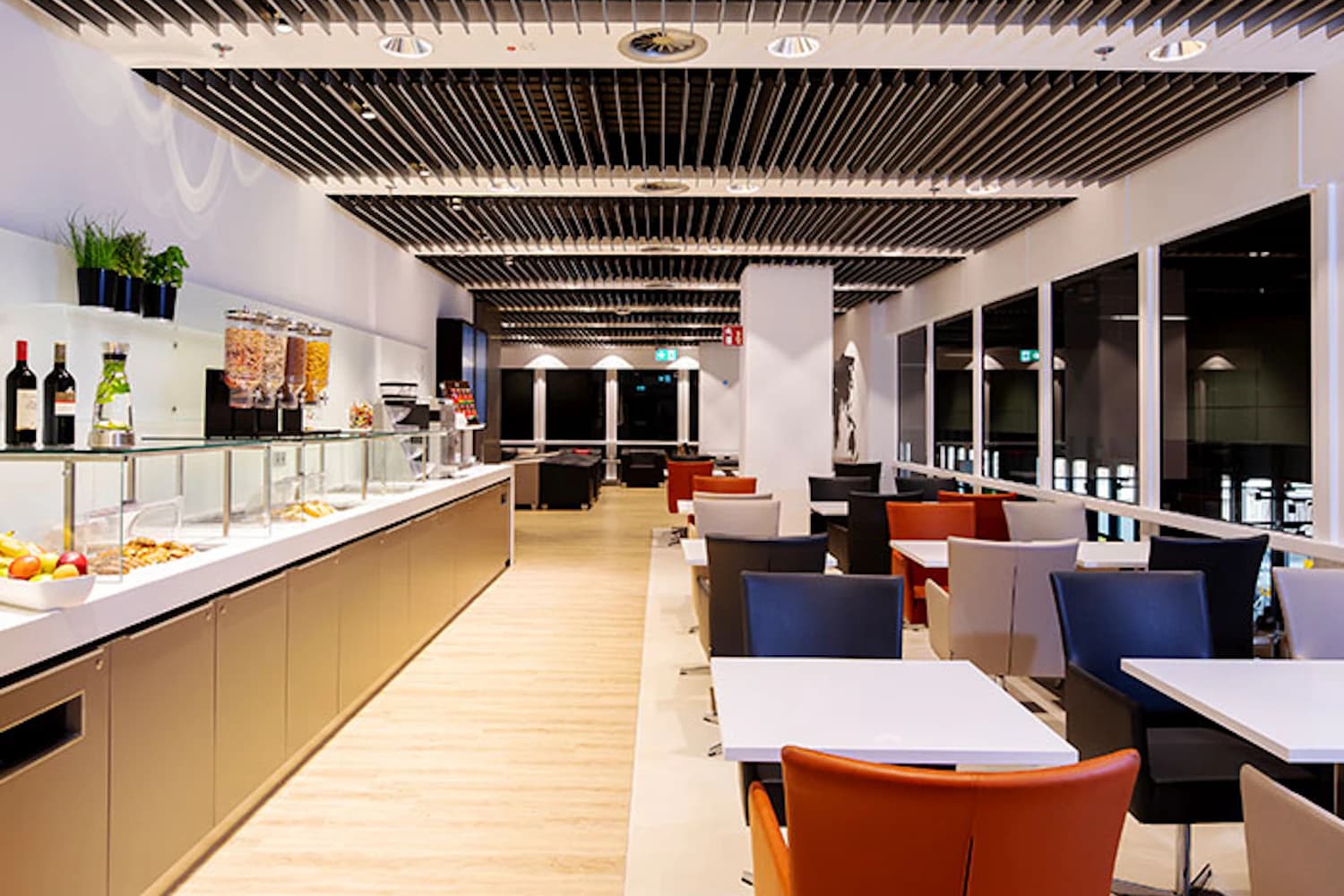 Helle Flughafenlounge mit Buffet links, Tischen und bunten Sesseln rechts sowie großen Fenstern.