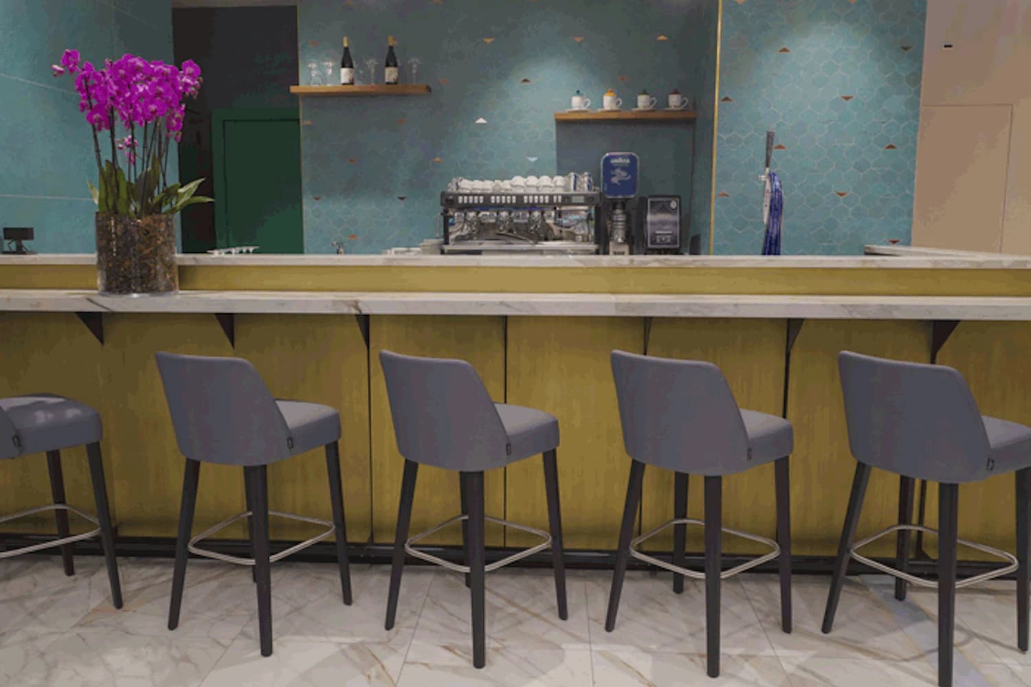 Bar mit marmorierter Arbeitsfläche, fünf graue Barhocker, Espressomaschine, Regale und violette Orchidee links.