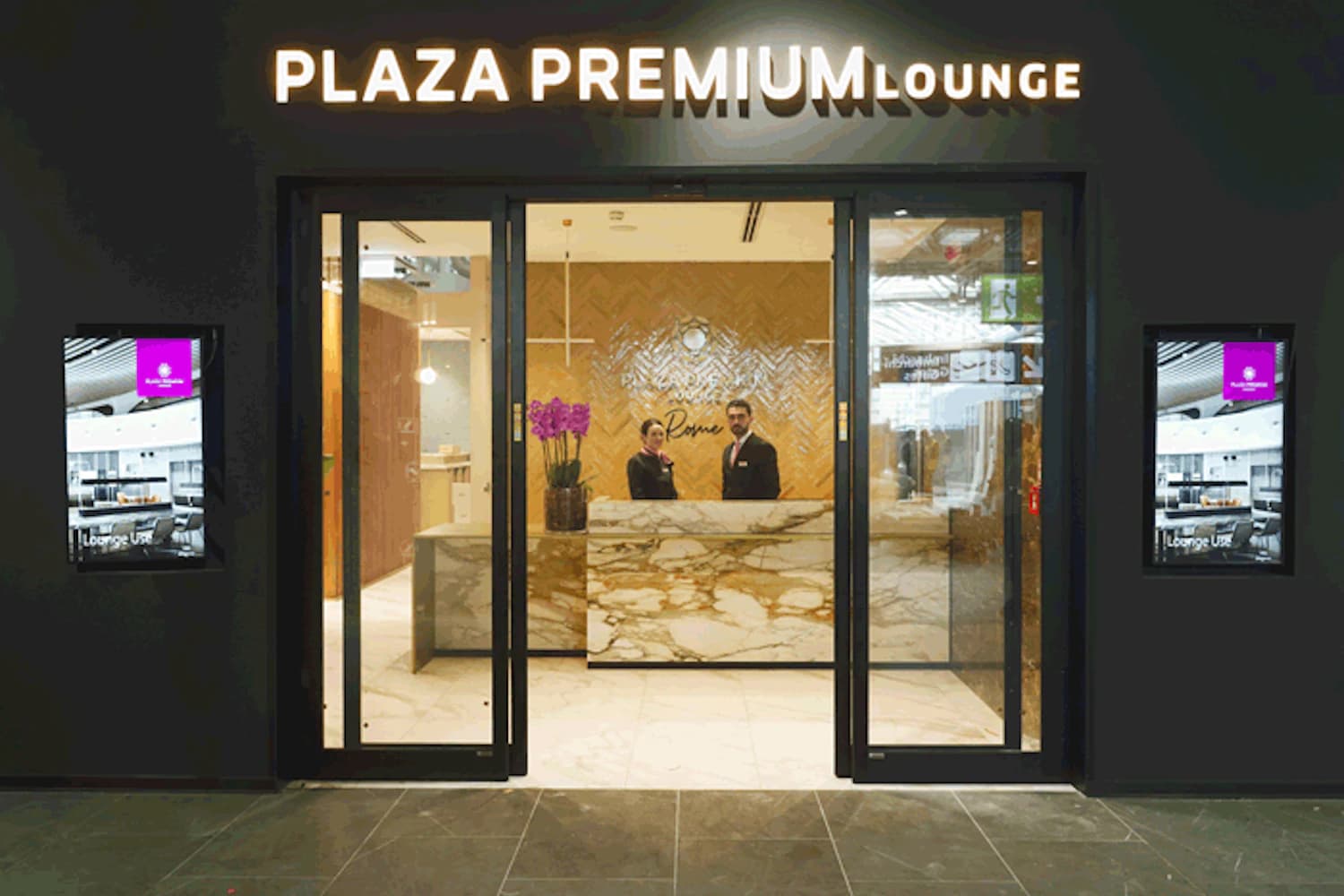 Eingang der Plaza Premium Lounge in Rom-Fiumicino: Glastüren, beleuchteter Schriftzug, Marmortresen und zwei Mitarbeitende.