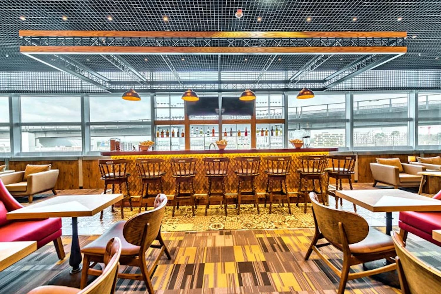 Loungebar mit Holztheke, Reihe Barhocker, hängenden Lampen und Sitzgruppen vor großen Fenstern.