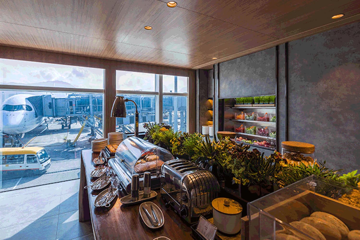 Buffetbereich einer Flughafenlounge mit Theke, Toastgeräten, Speisen und Fensterblick auf ein Flugzeug am Gate.