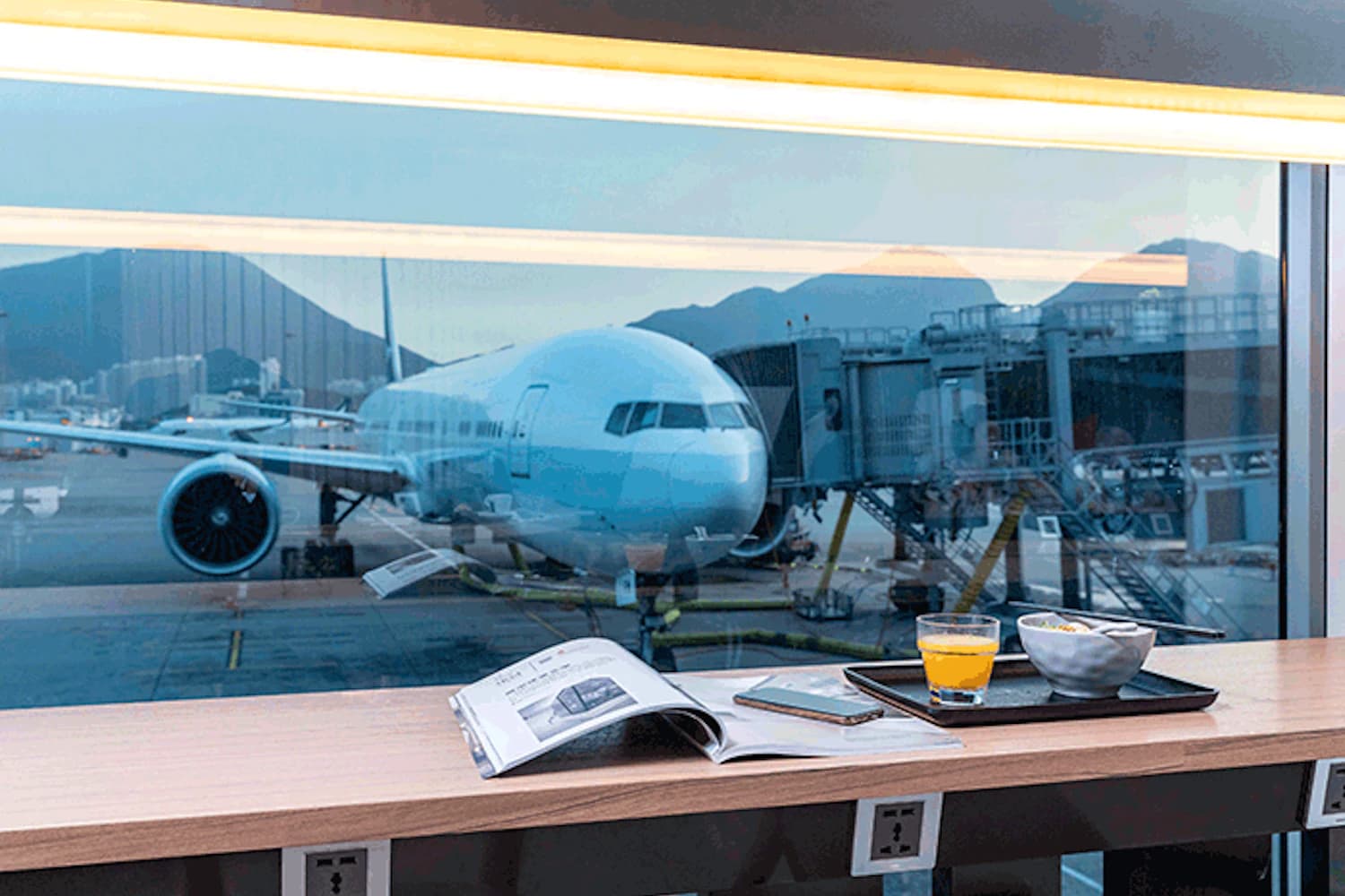 Fensterplatz in Flughafenlounge mit Magazin, Handy, Getränk und Schale; Flugzeug am Gate.