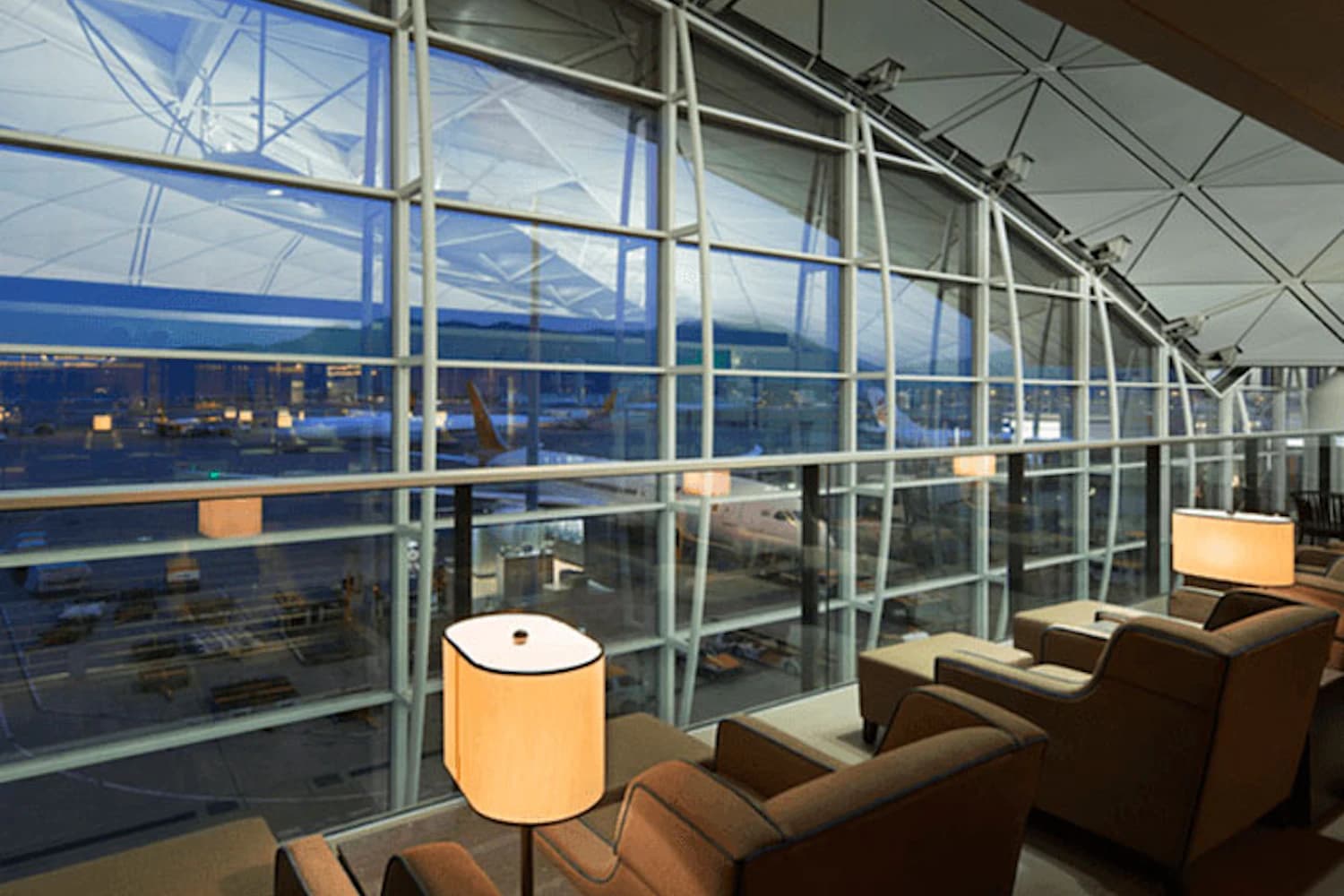 Reihen bequemer Sessel und Stehlampen vor großen Panoramafenstern mit Blick auf Flugzeug und Rollfeld.