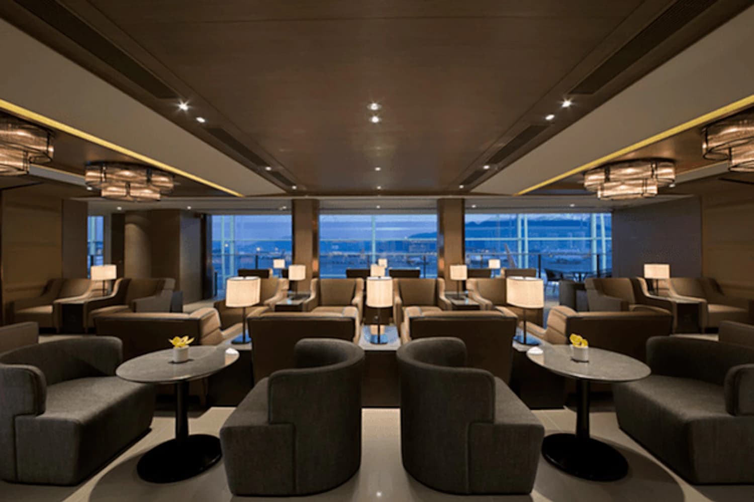 Lounge mit Reihen beiger und grauer Sessel, kleinen Tischen und Lampen vor großen Fenstern zum Flughafenvorfeld.