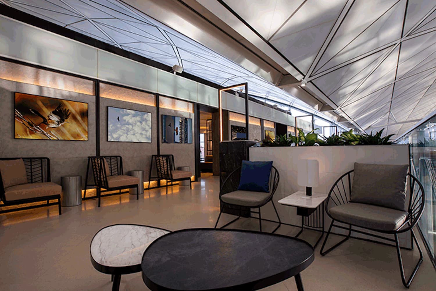 Lounge mit Sitzgruppen, niedrigen Tischen, Wandbildern, Pflanzen und hoher geometrischer Decke im Flughafenterminal.