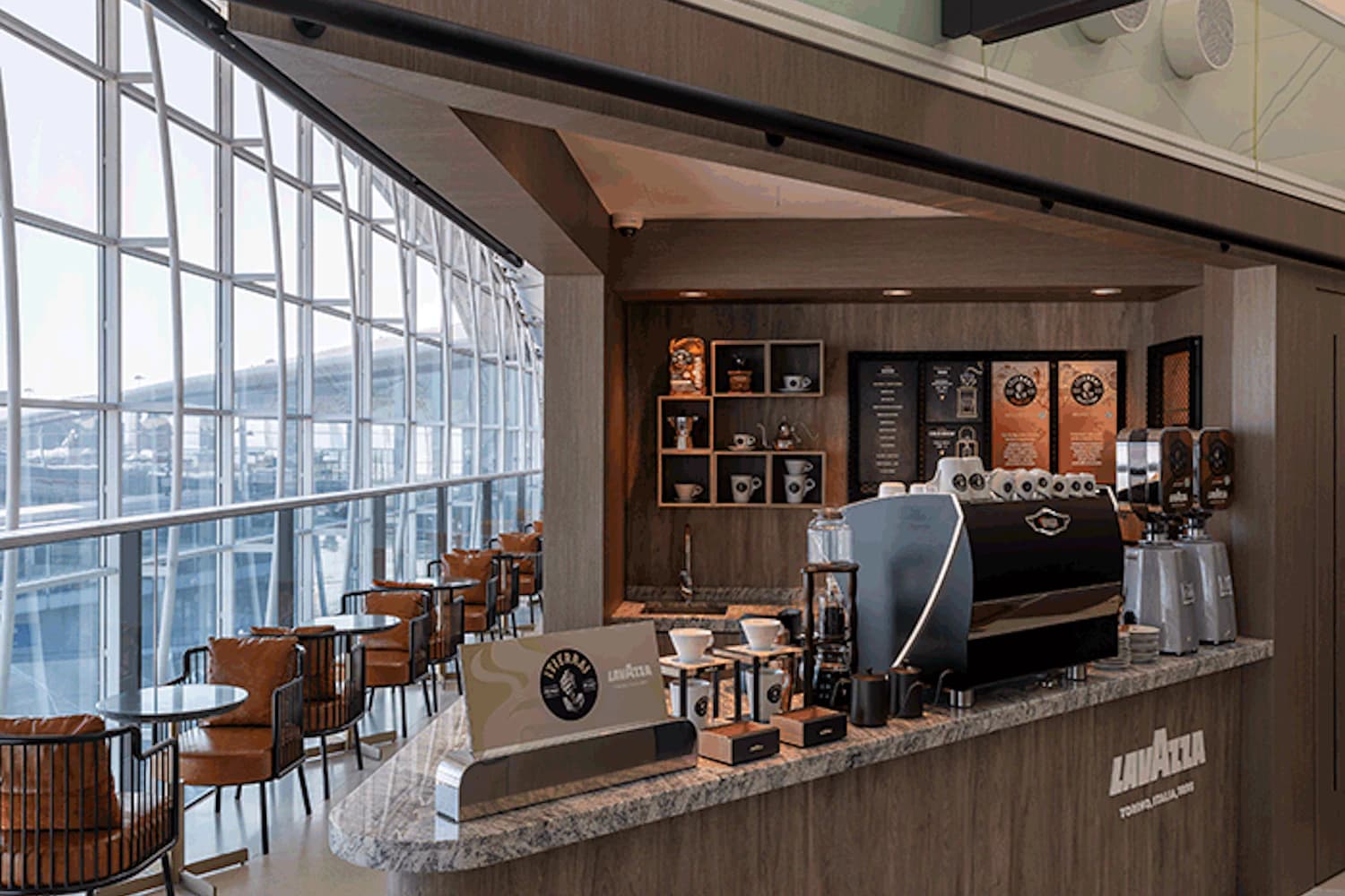 Kaffeebar mit Espressomaschine und Theke vor großer Fensterfront, Reihen von Sitzplätzen in Flughafenlounge.