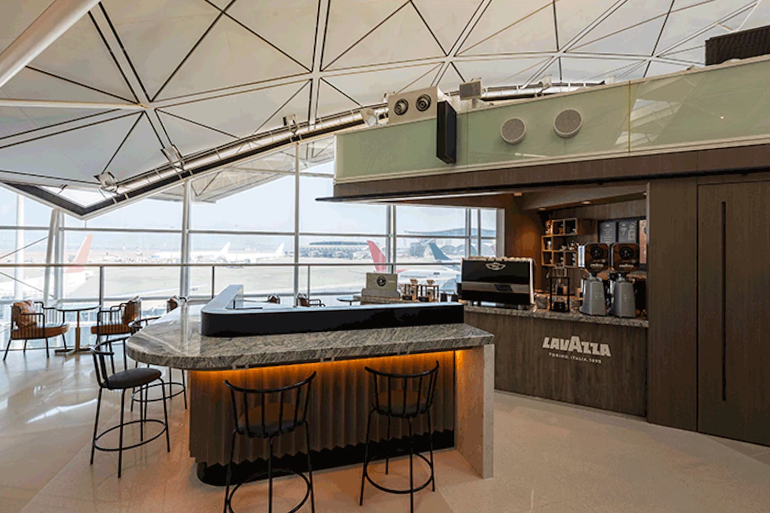 Offene Flughafenlounge mit Marmortheke, Barhockern, Lavazza-Kaffeezeile und großen Fensterfronten mit Flugzeugen außen.