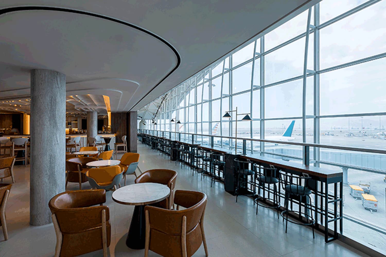 Weitläufige Flughafenlounge mit Tischen, Ledersesseln und großen Panoramafenstern mit Blick auf Rollfeld und Flugzeughecks.