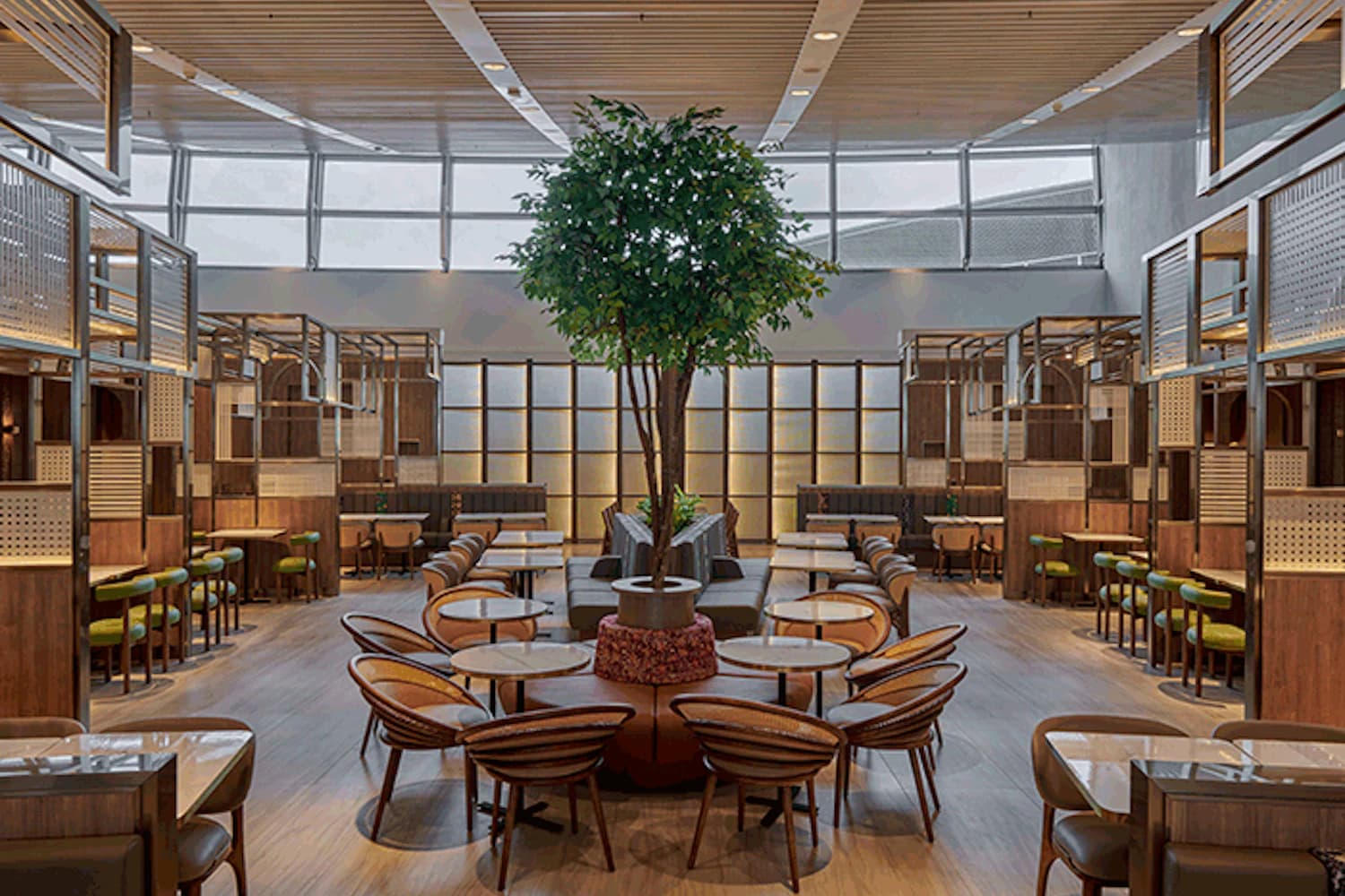 Offene Lounge mit mittigem Baum, runden Tischen, Holzstühlen und Sitznischen unter großem Fensterdach.