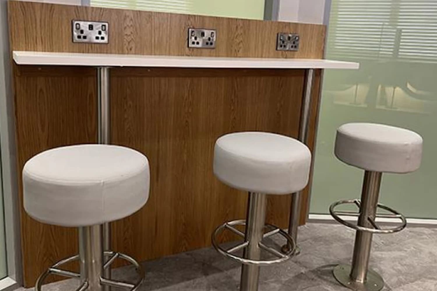 Drei runde weiße Hocker vor einer Holztheke mit Steckdosen in einer Flughafenlounge.