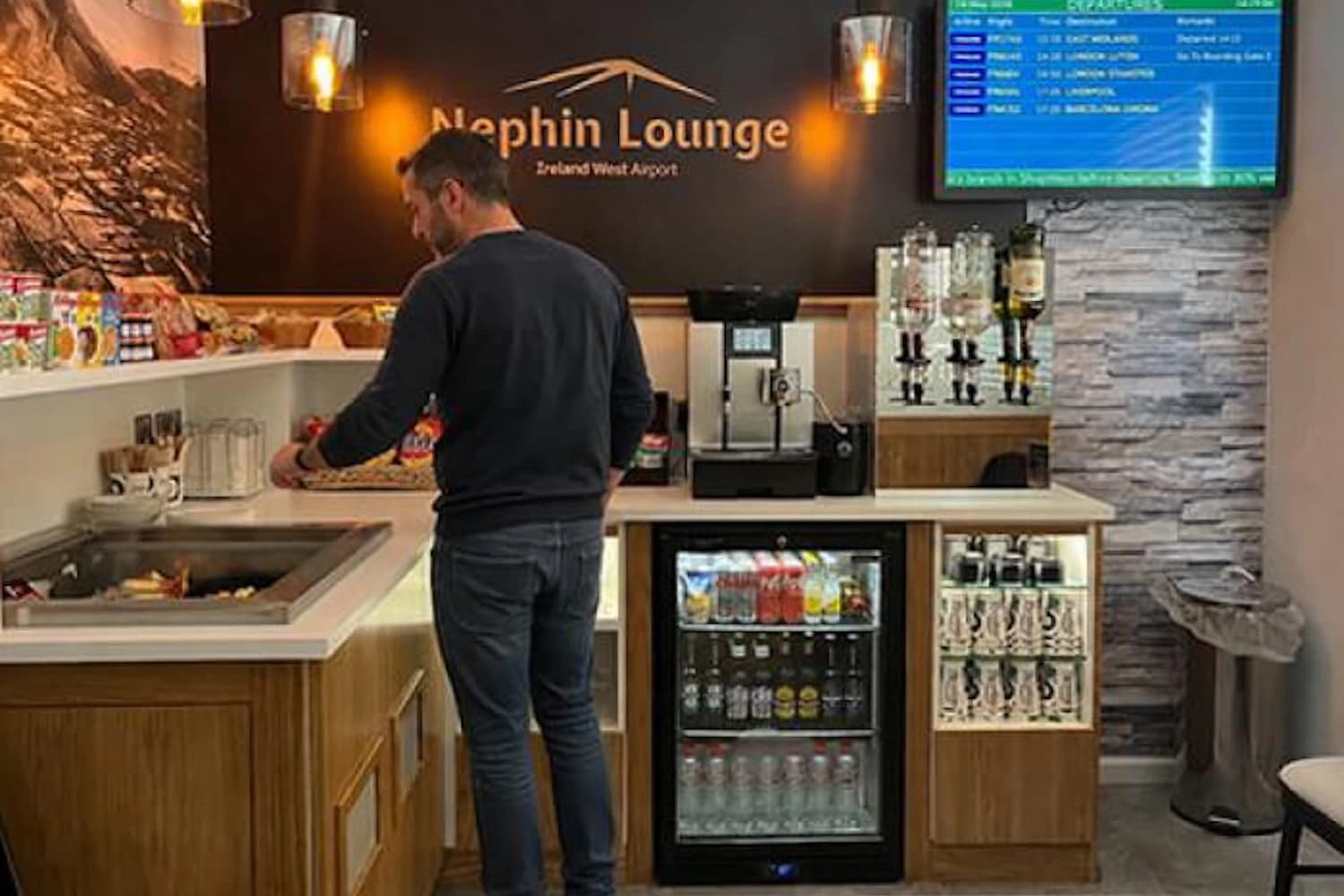Mann am Selbstbedienungsbuffet der Nephin Lounge mit Getränkekühlschrank, Kaffeemaschine und Abflugtafel.