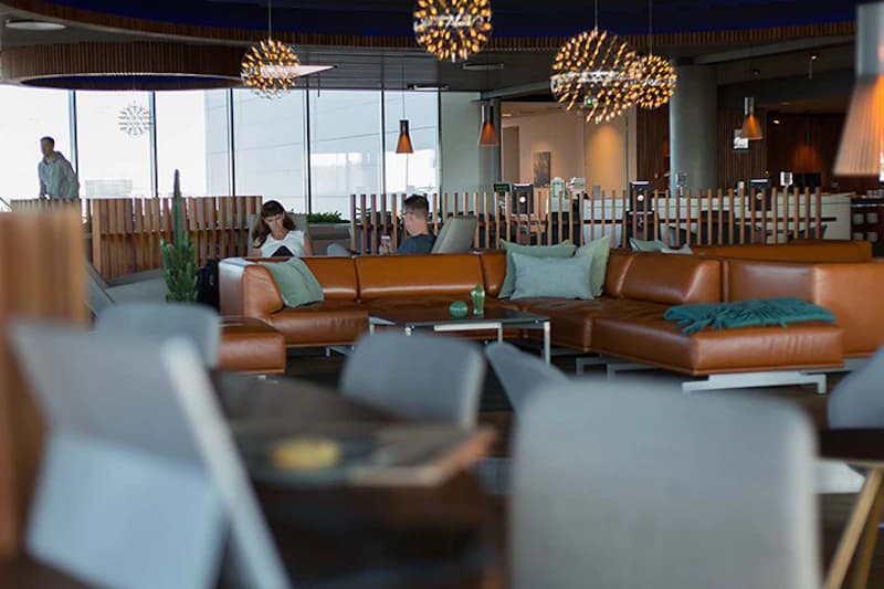 Lounge mit braunem Ledersofa, Tischen und hängenden Kugelleuchten, zwei Personen sitzen, große Fenster im Hintergrund.