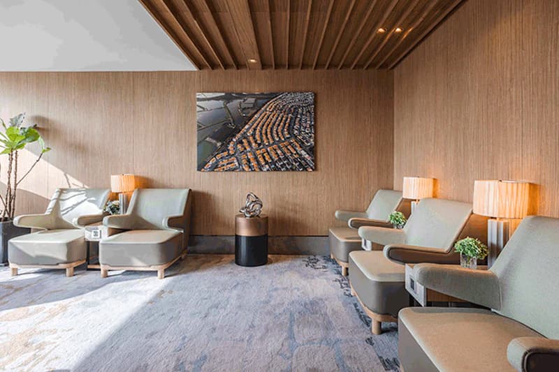 Helle Lounge mit mehreren Sesseln, Beistelltischen, Stehlampen, Teppichboden und Holzwand.