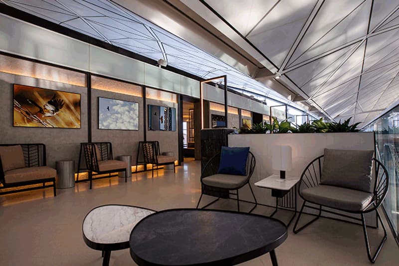 Lounge mit Sitzgruppen, niedrigen Tischen, Wandbildern, Pflanzen und hoher geometrischer Decke im Flughafenterminal.